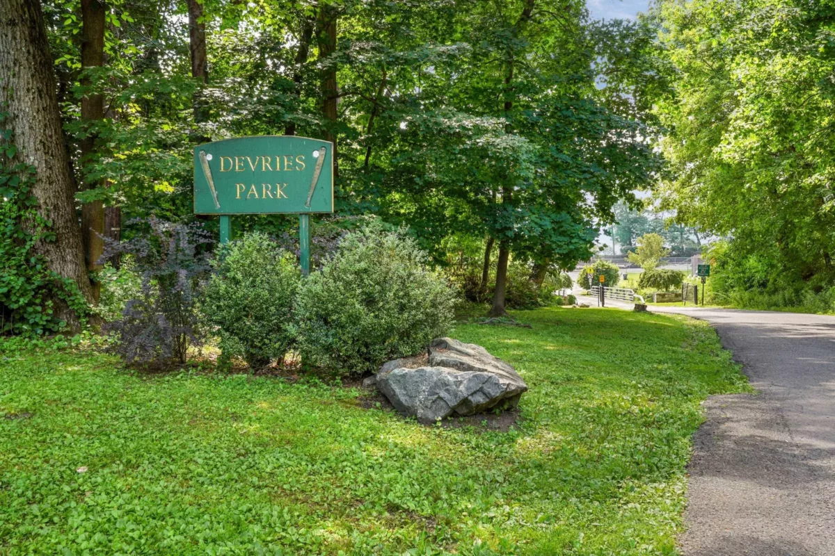 A pathway leading into a park next to trees, bushes, and a sign that says "DeVries Park".