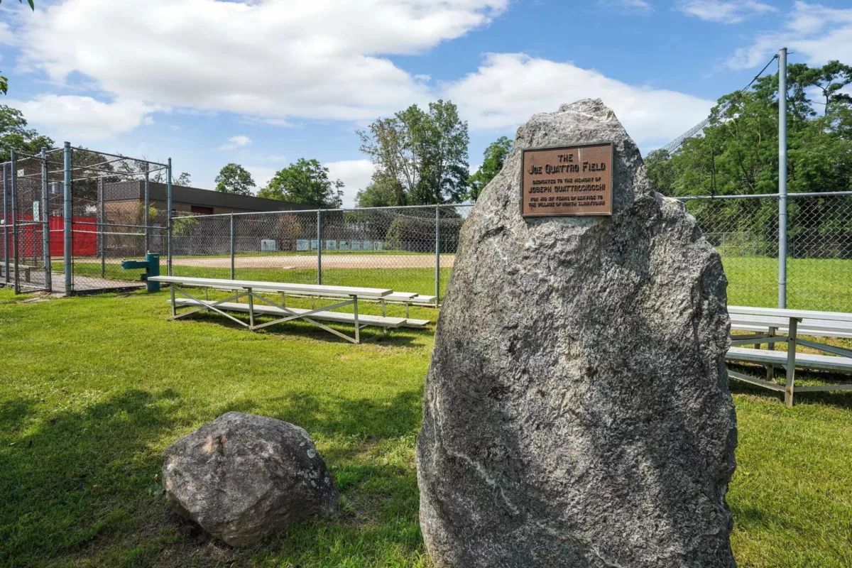 A large rock with a mounted plaque in front of a little league baseball field.