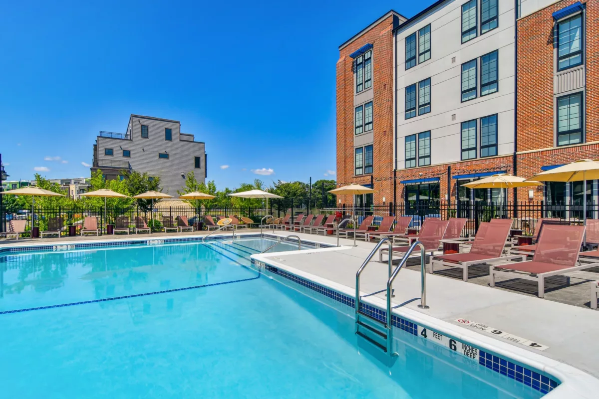 Outdoor pool area with lounge chairs and umbrellas beside a brick apartment building. The water is clear under a bright blue sky.