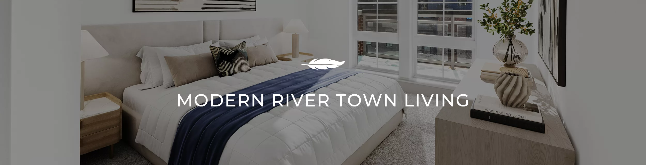 A modern bedroom with a large bed, two windows, and a light wood dresser with decor with the words "Modern River Town Living" across the center of the photo.